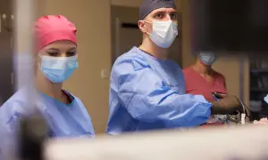 Chirurg w masce i fartuchu operacyjnym wykonuje zabieg, asystują mu dwie pielęgniarki.