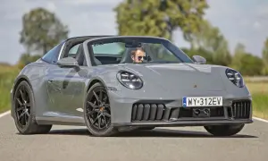 Szary Porsche 911 Carrera Targa z otwartym dachem, prowadzony przez mężczyznę w okularach przeciwsłonecznych, jedzie po asfaltowej drodze.