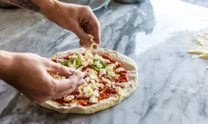 Hände belegen Pizza mit zerbröckeltem Käse auf Marmortisch.
