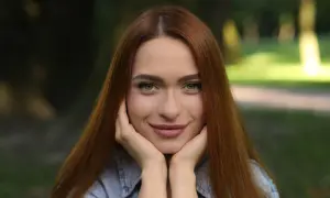 Uśmiechnięta szatynka z rudymi włosami opiera podbródek na dłoniach, patrząc w obiektyw.