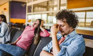 Erschöpfter Mann im Wartebereich, der sich die Augen reibt. Die Frau neben ihm wirkt ebenfalls müde. Gute **Jetlag Tipps** sind hier gefragt.