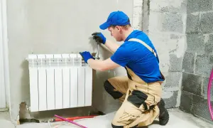 Instalator w niebieskiej czapce i ogrodniczkach pracuje przy grzejniku. Pokazuje, jak zdjąć grzejnik, używając klucza.