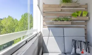 Przytulna loggia z szarą sofą, poduszką z liskiem i zielenią na drewnianej ścianie. Widok na drzewa przez przeszklony balkon.