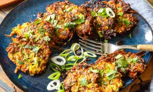 Knusprige Zucchini-Puffer, garniert mit frischen Kräutern und Frühlingszwiebeln, liegen auf einem blauen Teller. Eine Gabel liegt daneben.