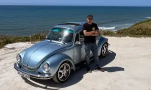 Man står bredvid en ljusblå Volkswagen Bubbla vid havet. En påminnelse om vem grundade Volkswagen och dess ikoniska design.
