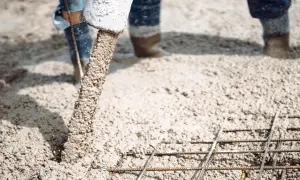 Beton na wylewkę płynie z rury, tworząc solidną podstawę dla budowy. Widać zbrojenie i nogi robotników w gumowcach.