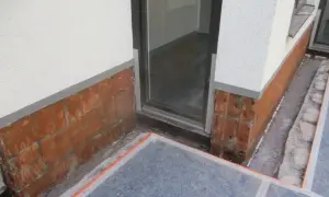 Rote Ziegelsteine am Sockel zeigen Anzeichen von aufsteigender Feuchtigkeit im Sockelbereich. Ein Fensterrahmen ist sichtbar.