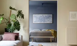 Ett mysigt blått vardagsrum med en stor Monstera och en soffa. Genom en öppning syns ett sovrum med en konstnärlig tavla.