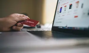 Mano sosteniendo tarjeta roja para pagar en plataformas de compra online. Pantalla de portátil muestra ofertas.