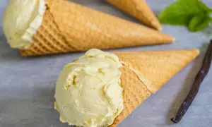 Drei Waffelkegel mit cremigem Vanilleeis, frisch aus der Eismaschine. Eine Vanilleschote und Minze liegen daneben.