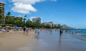 Słoneczny dzień na plaży Waikiki, z turystami kąpiącymi się w oceanie i odpoczywającymi na piasku, w tle palmy i hotele.