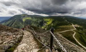 Kamienista ścieżka w górach, z łańcuchami zabezpieczającymi. Turyści wędrują, a krajobraz zachwyca zielenią i skalistymi zboczami. Pogoda groźna, chmury zapowiadają burzę, ale widok jest wart każdego kroku, jakbyśmy zbierali siły na ile z kopy na śnieżkę.