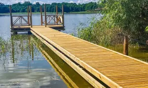 Drewniany pomost nad jeziorem zaprasza do spaceru. Woda odbija błękitne niebo i zielone drzewa.