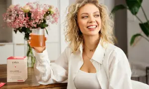 Uśmiechnięta kobieta z kręconymi blond włosami trzyma szklankę napoju. Obok stoi pudełko z napisem "Collagen Your Skin", sugerujące kolagen hydrolizowany.