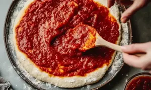 Händer sprider hemlagad egen pizzasås på en pizzabotten.