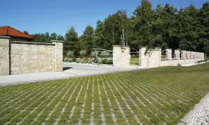 Piękny, kamienny mur i brama wjazdowa, a przed nimi zielona przestrzeń z ażurami na drogę.