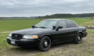 Svart Ford Crown Victoria, en robust bil som står parkerad på en grusväg med gröna fält och skog i bakgrunden.
