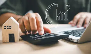 Person berechnet Miete unter Mietspiegel für das Finanzamt. Hausmodell, Rechner und Laptop im Fokus.