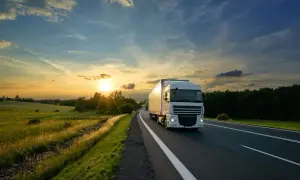 Un camion blanc roule sur une route au coucher du soleil, traversant une campagne verdoyante. C'est le quotidien de ceux qui travaillent sur la route.