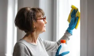 Mujer con gafas limpiando una ventana con un paño amarillo y guantes azules. La limpieza de exteriores es importante para mantener el hogar reluciente.