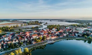 Poranna mgła nad rzeką otula malownicze miasteczko Sieraków Wielkopolski. Dachy domów lśnią w słońcu, a jezioro odbija błękit nieba.