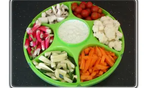 Plateau de crudités coloré : radis, champignons, tomates cerises, chou-fleur, courgettes et carottes, avec une sauce blanche au centre.