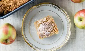 Pyszna szarlotka na talerzyku, posypana cukrem pudrem. Obok świeże jabłka. Idealny przepis na domowe ciasto.