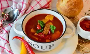 Miska gulaszowej zupy z mięsem, ziemniakami i papryczkami chili, obok chleb i przyprawy.
