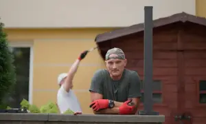 Mężczyzna w czapce i rękawiczkach opiera się o mur. W tle inny mężczyzna maluje drewnianą szopę. Jak zgłosić się do polowania na ogród?