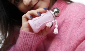 Delina Exclusif od Parfums de Marly, różowy flakonik z ozdobnymi frędzelkami, trzymany w dłoni na tle pudrowego swetra.
