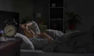A person lies awake in bed, hand on forehead, experiencing insomnia after surgery. An alarm clock sits on the nightstand.