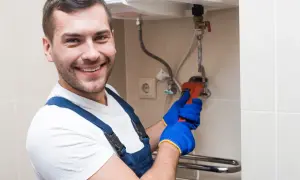 Uśmiechnięty hydraulik w niebieskim kombinezonie i rękawicach naprawia bojler. Dowiedz się, ile czasu grzeje się woda w bojlerze elektrycznym.