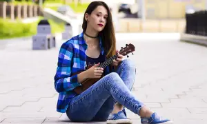 Dziewczyna w kratę i jeansach pokazuje, jak trzymać ukulele, siedząc na chodniku.
