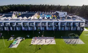 Widok z lotu ptaka na kompleks domków i budynków mieszkalnych z basenem i placem zabaw, na trawniku boisko do siatkówki i stoły do ping-ponga.