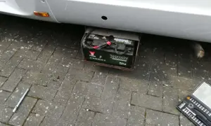 Emplacement batterie Ford Transit : une batterie Yuasa "Active Leisure & Marine" est visible sous le véhicule, dans un support métallique. Une clé à douille et une boîte à outils sont au sol.