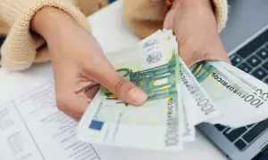 Mani che contano banconote da 100 euro, forse per la divisione conto corrente eredi. Documenti e laptop sullo sfondo.