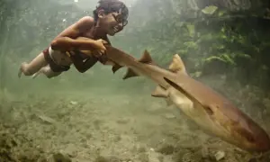 Un bambino del popolo Bajau, con occhiali da sub, nuota sorridendo accanto a uno squalo.