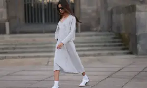 Elegancka sukienka i sportowe buty tworzą stylowy kontrast. Kobieta w szarym komplecie i białych sneakersach idzie ulicą.