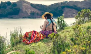 Frau mit Zöpfen und Hut sitzt am Seeufer, bunte Decke daneben. Ein Moment des bolivianischen Backpackings.