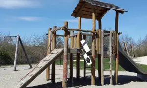 Drewniany plac zabaw ze zjeżdżalnią, drabinką i huśtawką na piaszczystym podłożu.
