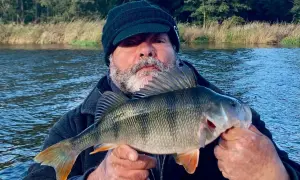 Ein Angler hält stolz einen schönen Barsch in den Händen. Das Wasser des Barther Bodden glitzert im Hintergrund.