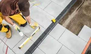 Operaio con martello di gomma e livella esegue la posa a secco di pavimenti esterni in lastre grigie, creando un bordo con pietre scure.