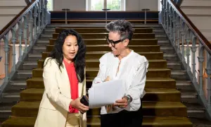 Två kvinnor diskuterar papper på en trappa. Den ena ger den andra en telefon, kanske för att hantera administrativa uppgifter.