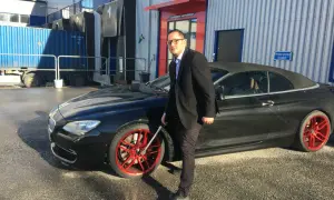 Man i kostym med glasögon vid en svart cabriolet med röda fälgar. Han håller i en fälgnyckel, redo att dra åt efterdra hjulbultar.