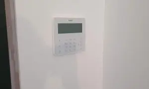 Biały panel alarmowy z klawiaturą numeryczną i ekranem na białej ścianie.