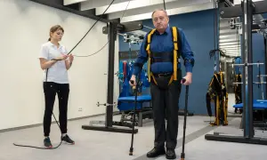 Starszy mężczyzna w specjalistycznej uprzęży i z kulami ćwiczy pod okiem fizjoterapeutki. To kluczowy etap rehabilitacji neurologicznej.
