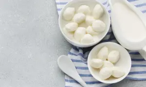Dwa białe naczynia z serkiem fior di latte, jedno z kulkami w zalewie, obok dzbanek z mlekiem i łyżka na tle z niebiesko-białym pasem.