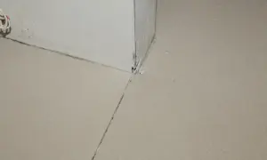 Uszkodzona dylatacja posadzki w rogu pomieszczenia, widoczne pęknięcia i ubytki materiału.