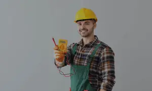 Uśmiechnięty elektryk w żółtym kasku i ogrodniczkach trzyma miernik. Dowiedz się, ile kosztuje przegląd instalacji elektrycznej.