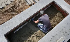 Budowlaniec wyrównuje świeży beton w wykopie. Widać ślady opon na błotnistym gruncie obok.
